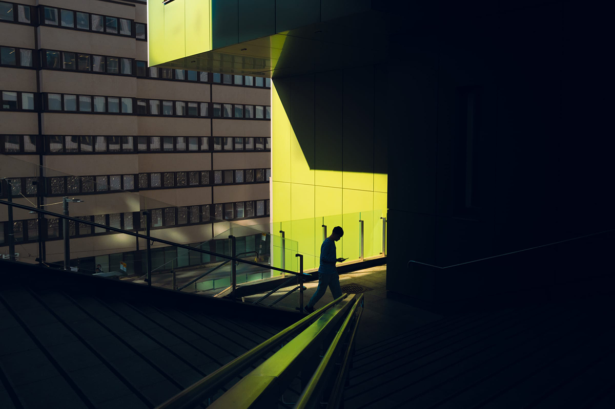 A man in a striped shirt walks through the last strip of low sun between a glowing yellow panel wall and deep shadow