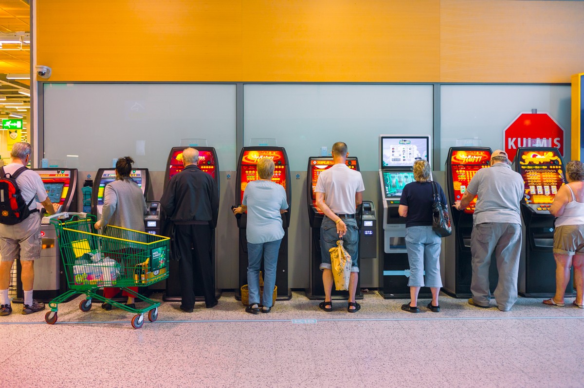 A row of people stand with their backs turned at nine glowing slot machines in a supermarket lobby, each absorbed in their own screen, one green shopping trolley parked behind them