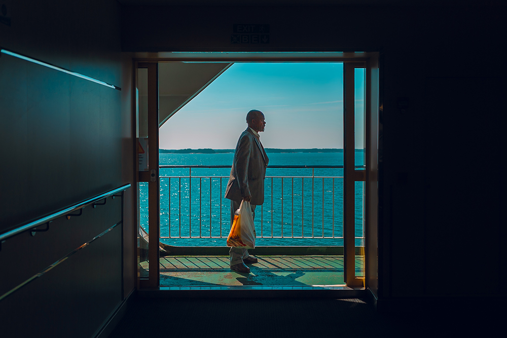 A man stands at the threshold of an open ferry door, looking out at the sea, suspended between the known and the unknown