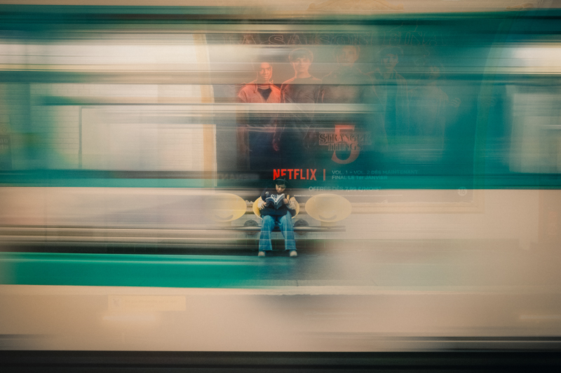 A lone figure sits perfectly still on a Paris metro platform, reading, while a train rushes past in motion blur around them, street photography by Loïc Sans