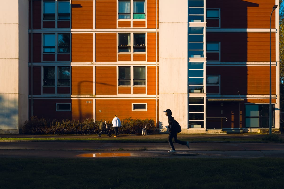 A silhouetted kid with a backpack runs through low morning sun in front of an orange apartment block